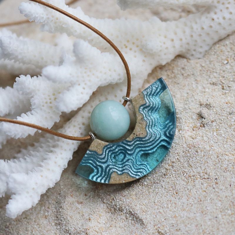 A half moon necklace with a blue bead in the dip of a reef made with sand and resin